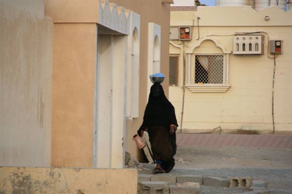 Woman in black veil walks past beige houses carrying a basin on her head