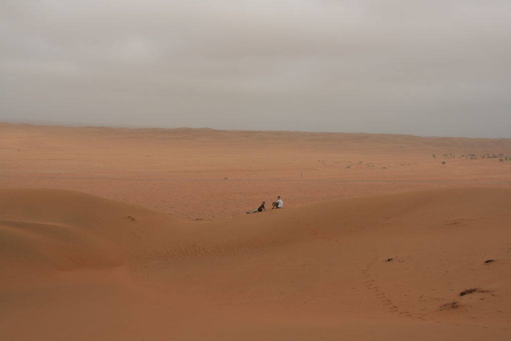 Two people sitting on an orange sand dune under a grey sky