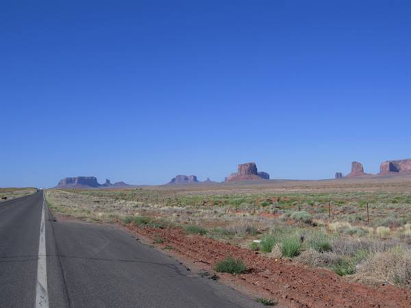 Asphalt road toward Monument Valley, red mesas among desert scrub under blue sky