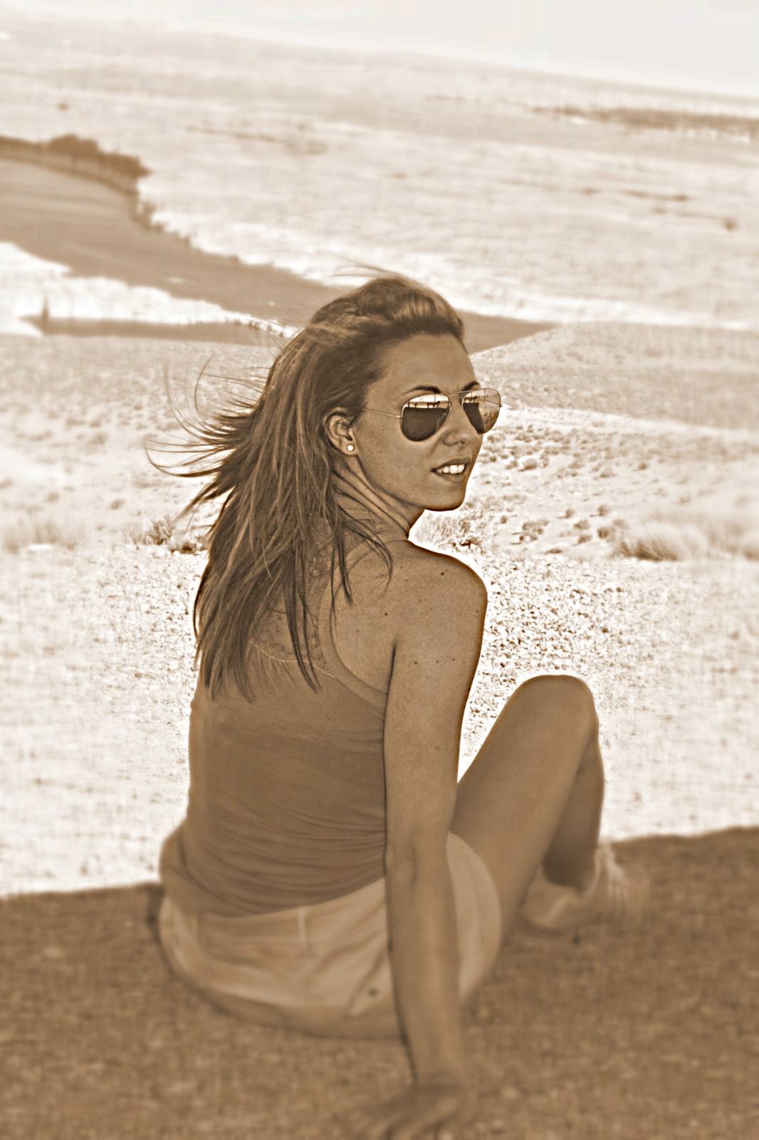 Woman in sunglasses sitting on the beach, hair blowing in the wind, sepia tone