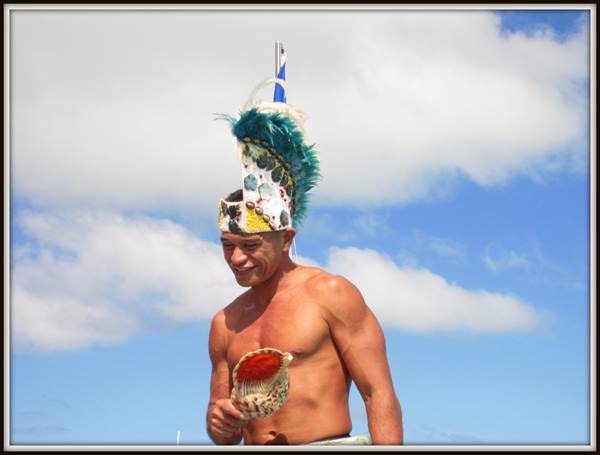 Shirtless man wearing green feather headdress blows a conch shell under blue sky