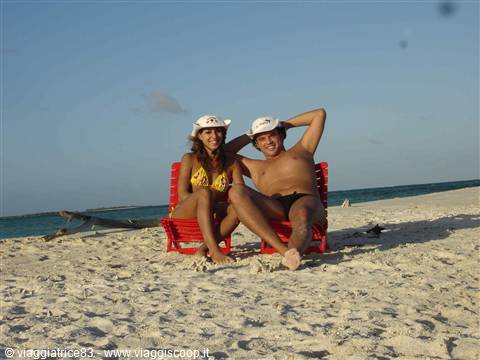Couple in white caps on red chairs over white sand facing turquoise sea