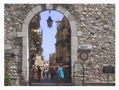 Woman in blue with open arms under Porta Messina, stone arch in Taormina