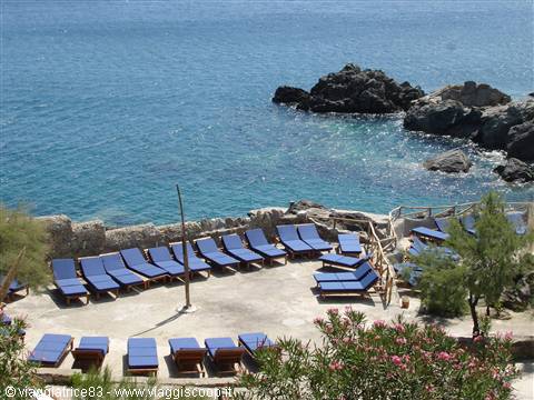 Blue sunbeds on a terrace over turquoise sea, black rocks, pink flowers