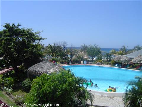 Blue swimming pool with thatched roofs, palm trees and people swimming by the sea
