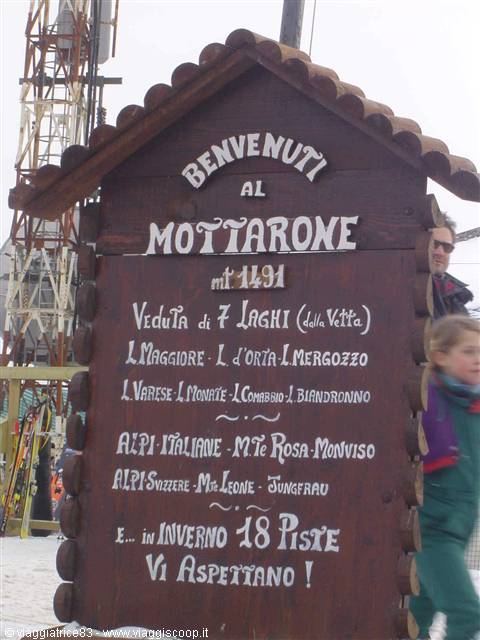 Wooden sign at Mottarone, 1491 meters, with skiers passing by in the snow