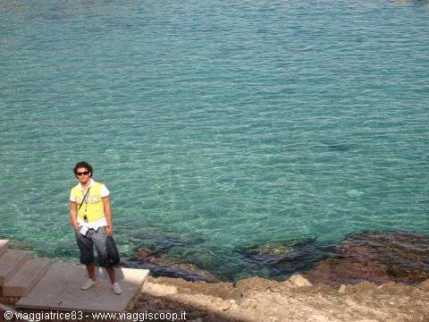 Woman in yellow shirt standing on rocks beside turquoise sea