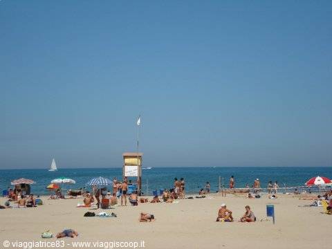 Crowded sandy beach with sunbathers, lifeguard tower, sailboats on calm sea