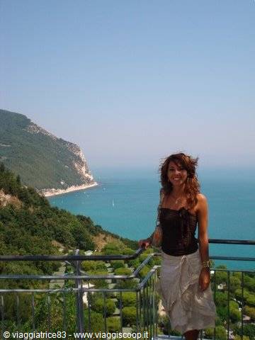 Smiling woman leaning on a metal railing, turquoise sea and Monte Conero coast behind