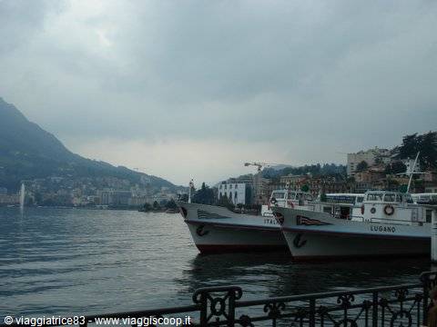 Ferry boats moored along Lugano lakefront, grey sky, mountains and fountain on the lake