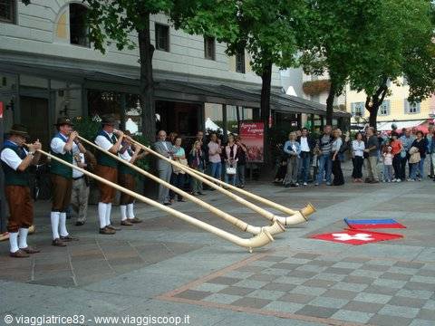 Musicians in traditional costume play alphorns on a town square crowd