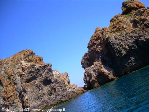 Reddish rock cliffs above turquoise water, seen from the sea under blue sky