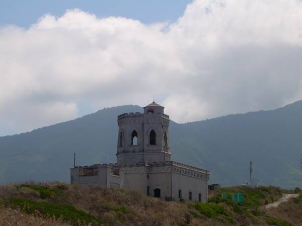 White crenellated stone tower on rocky hill, mountains and clouds behind