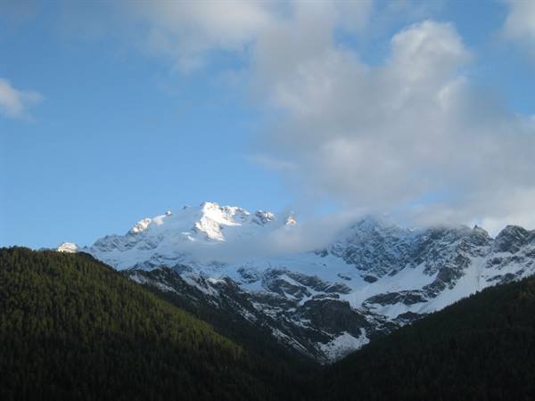 Snow-capped peaks lit by dawn above dark forests, clouds on blue sky