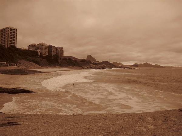 Waves washing Ipanema beach with high-rises and Dois Irmãos peak under cloudy sky