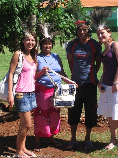 Four smiling people wearing feather headdresses, one showing a woven bag, sunny lawn