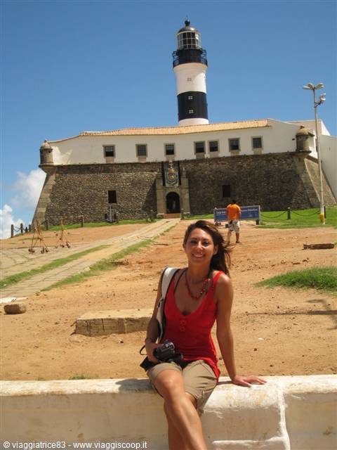Woman in red sitting on a wall in front of Farol da Barra, Salvador