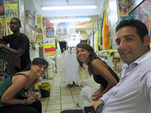 Smiling tourists in a souvenir shop with paintings and t-shirts, vendor nearby