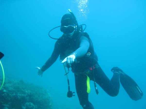 Scuba diver in black wetsuit pointing amid bubbles and coral reef