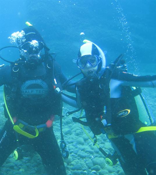 Two scuba divers in wetsuits pose underwater among stones