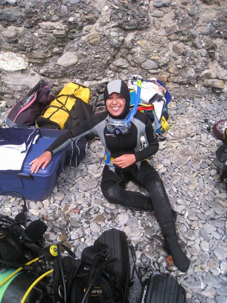 Diver in wetsuit sits on pebbles with mask and tanks