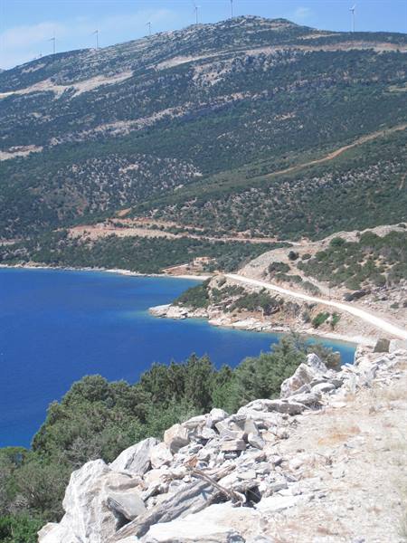 White gravel road along the coast, blue sea below green hills with wind turbines