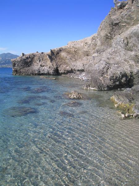 Clear shallow water over sand and rocks, grey volcanic cliff under blue sky