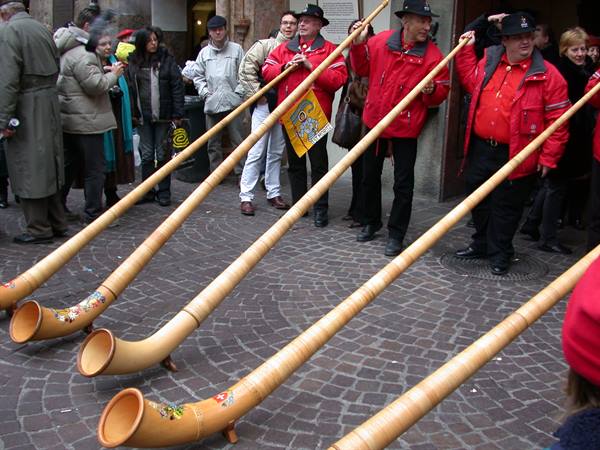 Musicians in red jackets play long wooden alphorns on cobblestones