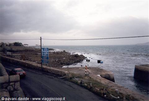 Car on rocky coastal road with pier and signs under cloudy sky
