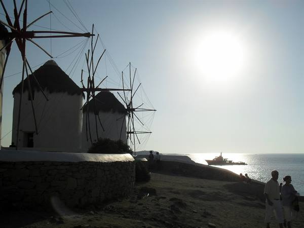 Mykonos white windmills against the sun, tourists and a ship offshore