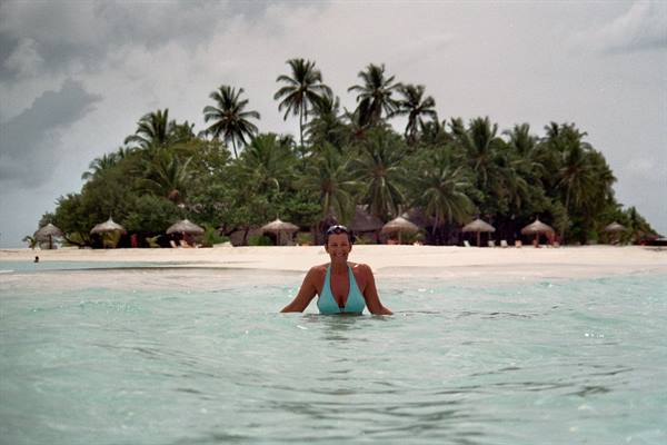 Woman in turquoise swimsuit wading by white beach with palm trees