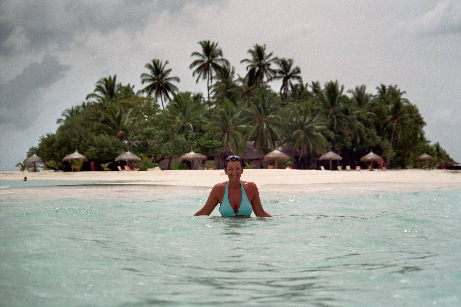 Woman in turquoise swimsuit wading by white beach with palm trees