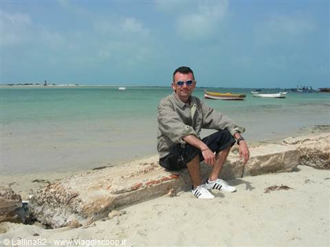 Man sitting on stone block on beach, colorful boats on turquoise sea