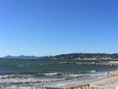 Sandy beach with waves, rocky breakwater and inhabited hills under clear blue sky