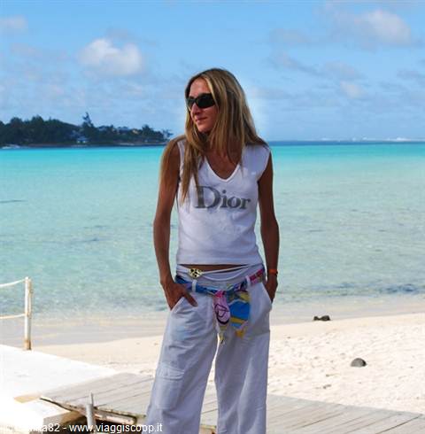 Blonde woman in sunglasses and Dior shirt on wooden boardwalk, turquoise lagoon