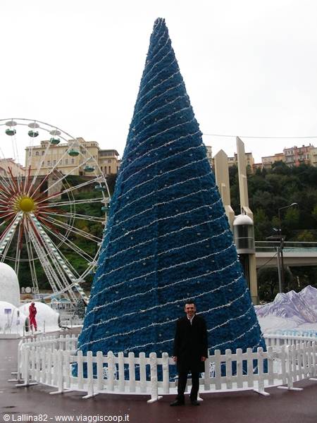 Man in black coat beside a blue Christmas tree, ferris wheel and hillside houses behind