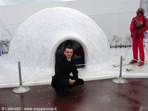 Man in black suit crouching before a snow igloo, skier in red suit nearby