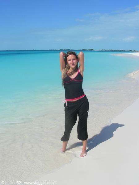 Woman in black raises her arms on white sand before turquoise water