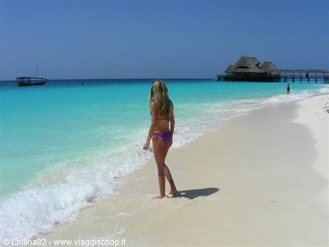 Woman in purple bikini walking on white sand near wooden pier and thatched bar