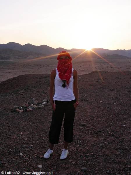 Woman with red keffiyeh covering her face in rocky desert at sunrise