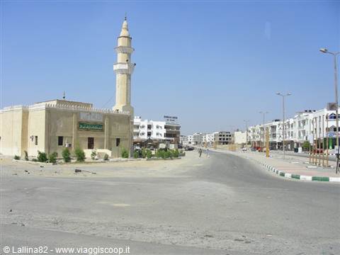 White mosque with minaret along sandy road, low buildings under blue sky