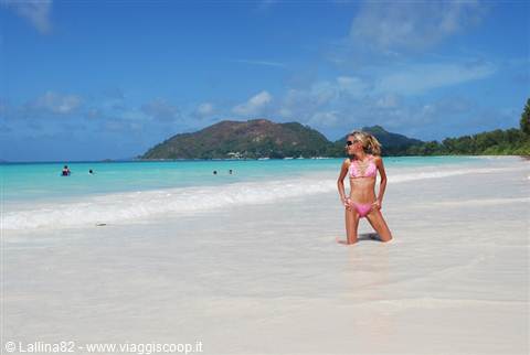 Woman in pink bikini kneeling on white sand, green hill and turquoise water