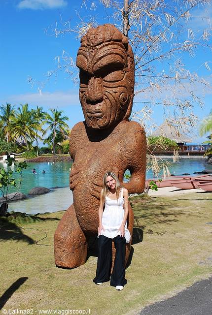 Tourist in white beside a giant red stone tiki statue, lagoon and palms