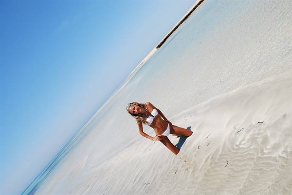 Woman in white bikini kneeling on pale sand under clear blue sky
