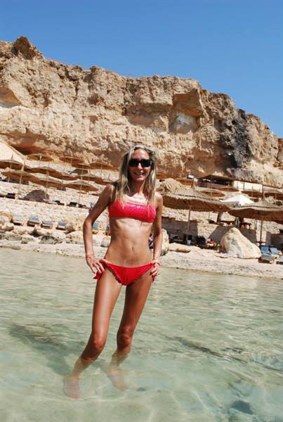 Woman in red bikini and sunglasses in clear water before sandy cliffs with beach umbrellas
