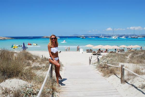 Woman in white bikini on wooden boardwalk leading to beach with umbrellas and boats