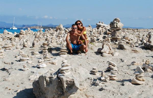 Couple crouching among stacked pebbles on white sand beach, sailboats offshore
