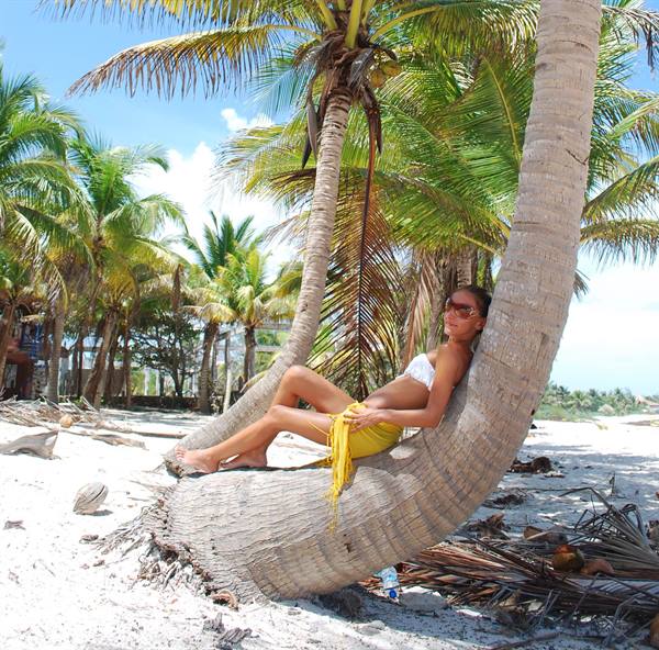 Woman reclining on a leaning palm tree over white sand among sunny palms