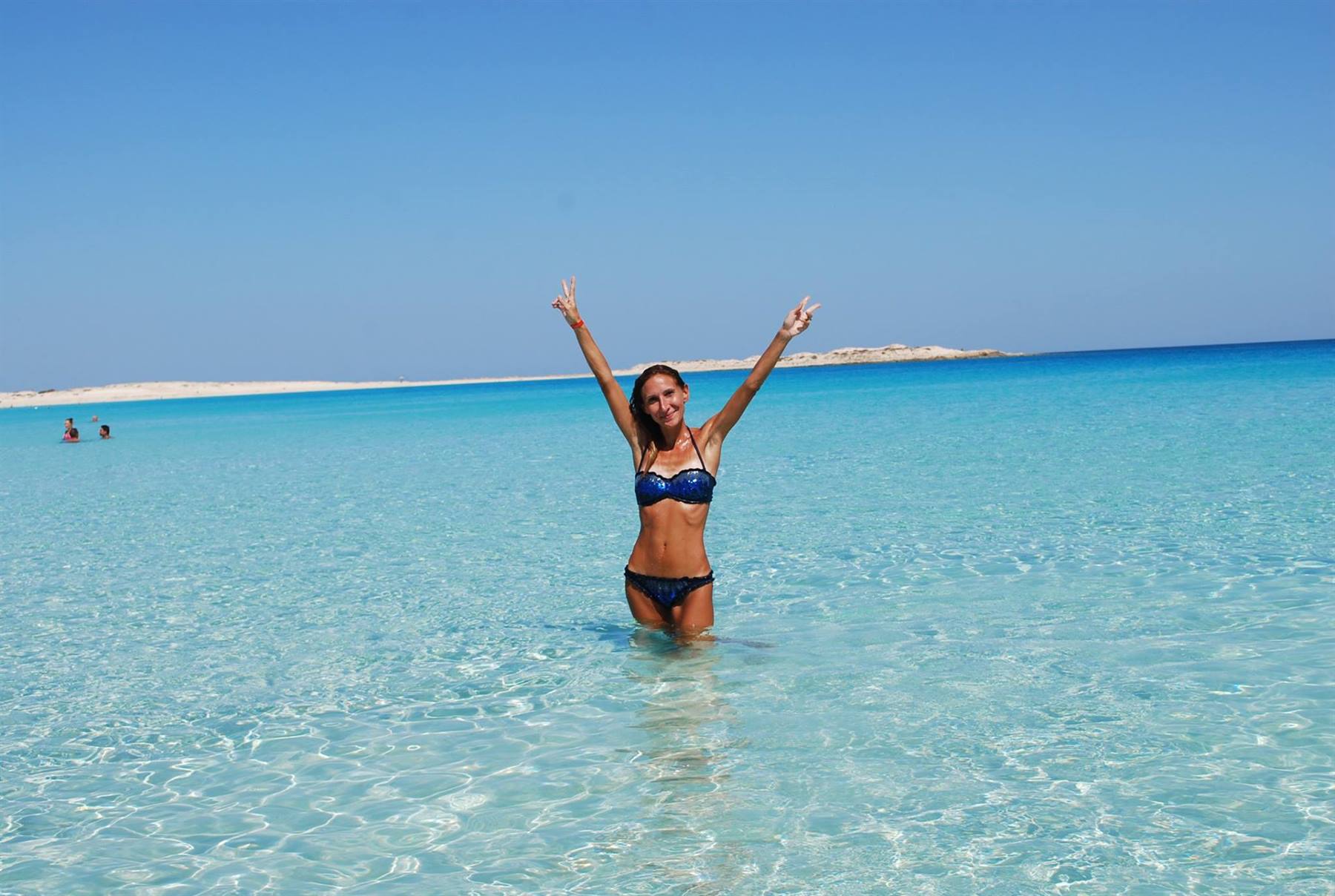 Donna in bikini braccia alzate nell'acqua turchese, spiaggia bianca sullo sfondo