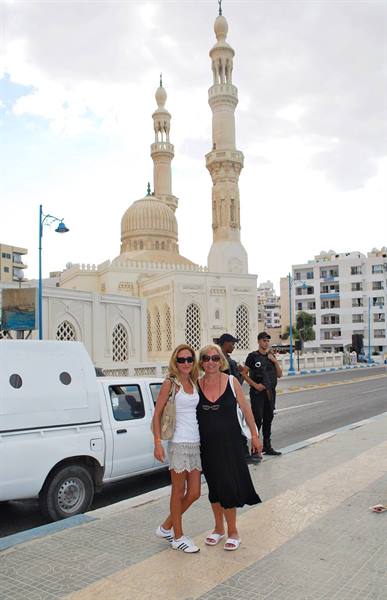 Two tourists pose on the street in front of a white mosque with minaret